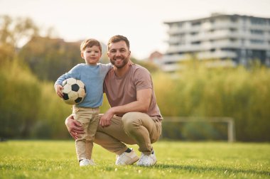 Oturuyor, futbol topu tutuyor. Mutlu baba ve oğul yazın sahada eğleniyorlar..