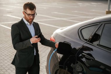 Şarj için mavi kablo hazır. Takım elbiseli bir işadamı dışarıda siyah arabasının yanında..