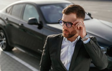 Gözlüklü, ön manzaralı. Takım elbiseli bir işadamı dışarıda siyah arabasının yanında..