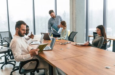 Masanın yanında oturuyor. Ofis çalışanlarından oluşan ekip içeride bir arada..