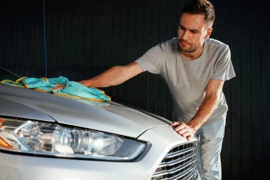 Adam kumaş kullanarak otomobilin yüzeyini cilalıyor. Adam dışarıda arabasıyla bekliyor..