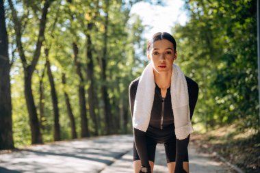 Boynunda beyaz havlu var. Genç, sıska kadın dışarıda siyah spor kıyafetleri giyiyor..