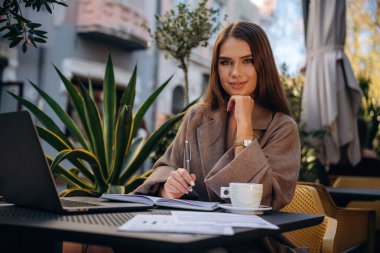 İçki ve belgelerle uzaktan çalışma. Güzel, genç bir bayan restoranın dışında, masanın yanında..