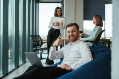 Yakışıklı adam dizüstü bilgisayarla oturuyor. Ofisteki insanlar koltuklarda fasulye torbalarıyla çalışıyorlar..