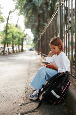 Akıllı telefon elinde, oturuyor. Sırt çantalı kız öğrenci dışarıda..