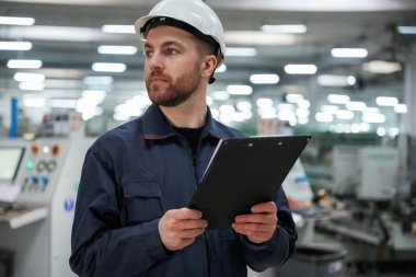 Uzağa bakıyor, not defterini tutuyor. Fabrika işçisi kasketli bir şekilde içeride..