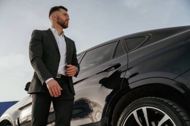 Doğal ışıklandırma, gündüz. Takım elbiseli bir işadamı dışarıda siyah arabasının yanında..