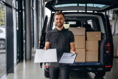 Kalem ve not defteri ellerinde. Teslimatçı ya da yükleyici elinde bir kutuyla arabanın yanında..