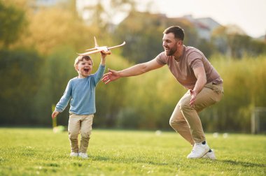 Günlük kıyafetlerle. Oyuncak uçakla koşuyor. Mutlu baba ve oğul yazın sahada eğleniyorlar..
