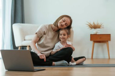 Genç anne ve kızı yoga minderinde oturup dizüstü bilgisayarı izliyorlar..