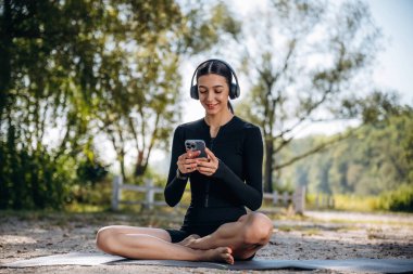Rahatlamış, yoga minderinde oturuyor, mola. Genç, sıska kadın dışarıda siyah spor kıyafetleri giyiyor..