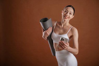 Akıllı telefon ve yoga minderi ellerinde. Güzel fitness kadını stüdyoda..