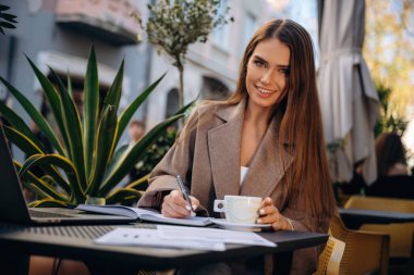 İçki ve belgelerle uzaktan çalışma. Güzel, genç bir bayan restoranın dışında, masanın yanında..