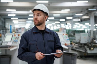 Uzağa bakıyor, not defterini tutuyor. Fabrika işçisi kasketli bir şekilde içeride..