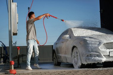Otomotiv temizleyicisi sabun ve köpüğü yüksek basınçlı çamaşır makinesiyle yıkıyor. Adam dışarıda arabasıyla bekliyor..