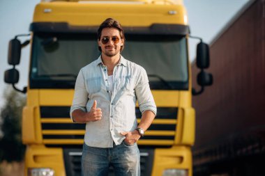 Göz giysisi, güneş gözlüklü havalı görünüm, ayakta durmak. Kamyon şoförü açık havada nakliye aracının yanında..