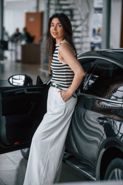 Aracın üzerinde duruyor ve yaslanıyor. Güzel genç bayan araba galerisinde yeni arabasıyla..