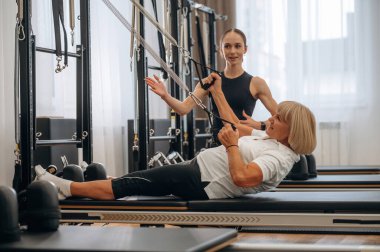 Eğitim ekipmanlarını kullanarak. Kadın eğitmen yaşlı kadının egzersiz yapmasına yardım ediyor..