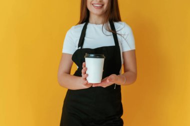 Siyah beyaz kahve işçisi üniforması, elinde bir bardak içki. Genç bayan sarı arka plana karşı..