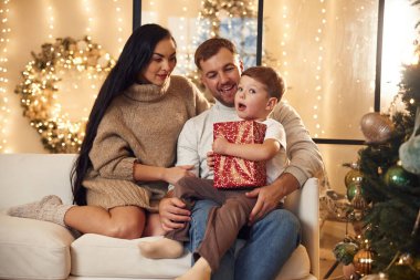 Kırmızı hediye kutusu bir çocuğun elinde. Mutlu bir aile yeni yılı evde kutluyor..