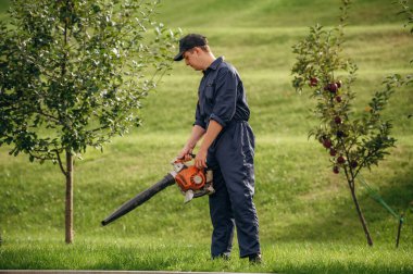 Yaprak üfleyici elinde, yolu temizliyor. Bahçede profesyonel bahçıvan var..