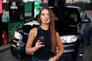 Elinde siyah bir bardak içki var. Benzin istasyonunda arabasıyla genç bir kadın..