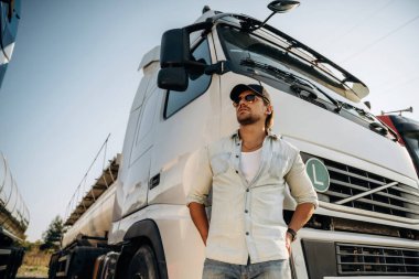 Aşağıdan bak, ayakta ve poz veriyor. Kamyon şoförü açık havada nakliye aracının yanında..