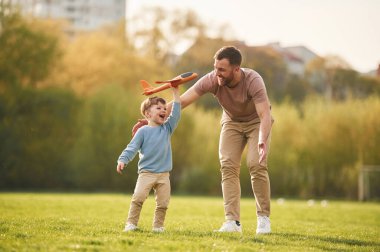 Günlük kıyafetlerle. Oyuncak uçakla koşuyor. Mutlu baba ve oğul yazın sahada eğleniyorlar..