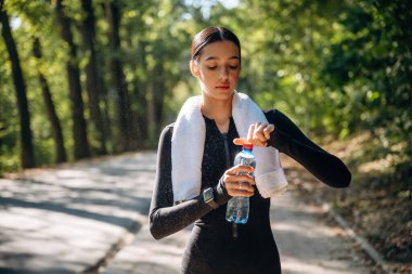 Boynunda beyaz havlu var. Genç, sıska kadın dışarıda siyah spor kıyafetleri giyiyor..