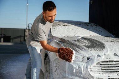 Otomotiv temizleyicisi sabun ve köpüğü yüksek basınçlı çamaşır makinesiyle yıkıyor. Adam dışarıda arabasıyla bekliyor..