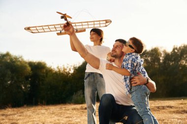 Oturuyorum, yandan bakıyorum. Baba, oğul ve kız çocukları sahada tahta oyuncak uçakla oynuyorlar..