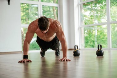 Pencerenin dışındaki yeşil ağaçlar. Güçlü kaslı adam spor salonunda çalışıyor..