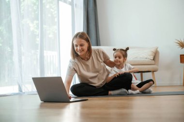 Laptop kullanıyorum. Küçük kızı olan genç bir kadın evde yoga yapıyor..