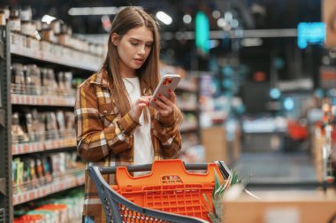 Akıllı telefonu kullanmak, ayakta durmak. Kadın süpermarkette alışveriş yapıyor..