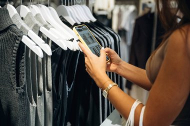 Akıllı telefon ellerde. Perakende dükkanında kıyafet seçen bir kadının yakın görüntüsü..