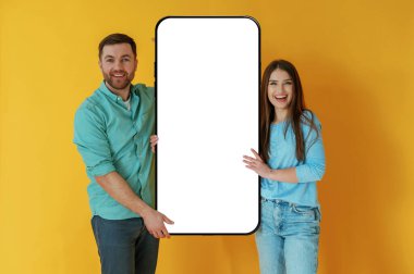 Kopyalama alanı için göster. Kadın ve erkek, sarı arka planda büyük bir akıllı telefonla duruyorlar..