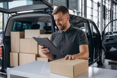 Yüz ifadesi olumlu, not defteri tutuyor. Teslimatçı ya da yükleyici elinde bir kutuyla arabanın yanında..