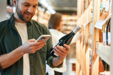 Akıllı telefon ve şişe bir adamın elinde. Erkek ve kadın dükkanda şarap seçiyorlar..