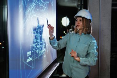 Beyaz kasketli. Kadın mimar projektör üzerine inşaat alanı planı gösteriyor.