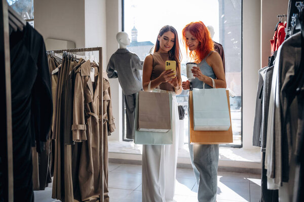 Some information in smartphone, reading. Two female friends are in the retail store, choosing clothes, shopping.