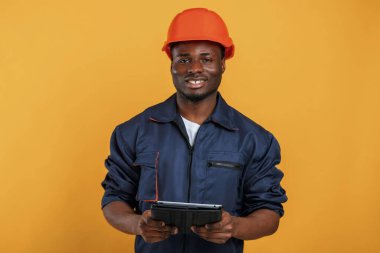 Kasketli ve tabletli inşaat işçisi. Yakışıklı siyah adam sarı arka plana karşı stüdyoda..