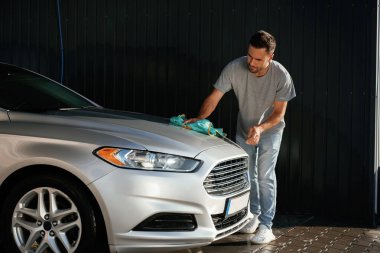 Adam kumaş kullanarak otomobilin yüzeyini cilalıyor. Adam dışarıda arabasıyla bekliyor..