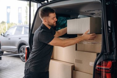 Paketlenmiş malı ellerinde tutuyorlar. Teslimatçı ya da yükleyici elinde bir kutuyla arabanın yanında..