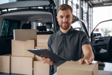 Yüz ifadesi olumlu, not defteri tutuyor. Teslimatçı ya da yükleyici elinde bir kutuyla arabanın yanında..