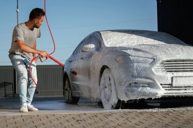 Otomotiv temizleyicisi sabun ve köpüğü yüksek basınçlı çamaşır makinesiyle yıkıyor. Adam dışarıda arabasıyla bekliyor..