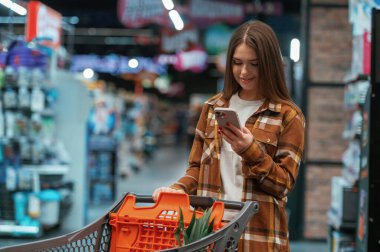 Akıllı telefonu kullanmak, ayakta durmak. Kadın süpermarkette alışveriş yapıyor..