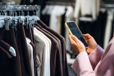 Akıllı telefondan bilgi okuyorum. Perakende dükkanında kıyafet seçen bir kadının yakın görüntüsü..