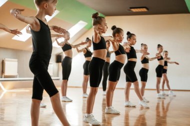 Eller yanda, ayakta. Bir grup bayan çocuk içeride spor egzersizleri yapıyor..