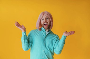 Yüz ifadeleri, el hareketleri. Pembe saçlı adam sarı arka planda poz veriyor..