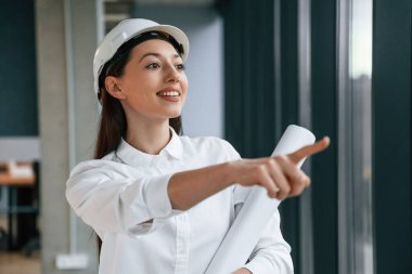 Parmaklarıyla kenarı işaret ediyor. Kasketli ve beyaz elbiseli kadın mimar içeride..